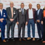 A smiling group, six men and one woman, standing in front of a banner printed with the Prisma and Clemson logos at the Center for Human Factors in Health Care launch