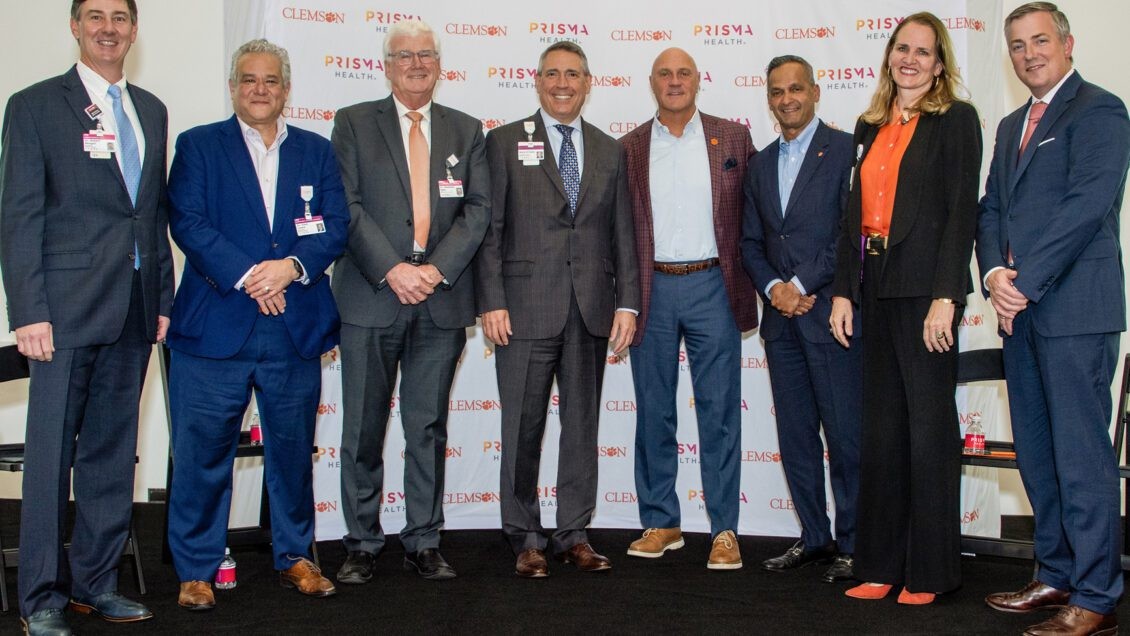 A smiling group, six men and one woman, standing in front of a banner printed with the Prisma and Clemson logos at the Center for Human Factors in Health Care launch